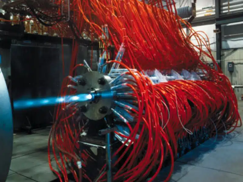 High-pressure rocket engine injector test with multiple red fluid lines and blue combustion jet, conducted in advanced aerospace research facility.