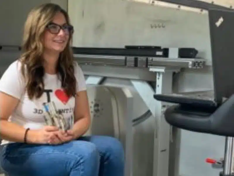 Melanie Lang in 'I Love 3D Printing' shirt holding metal parts inside industrial additive manufacturing lab